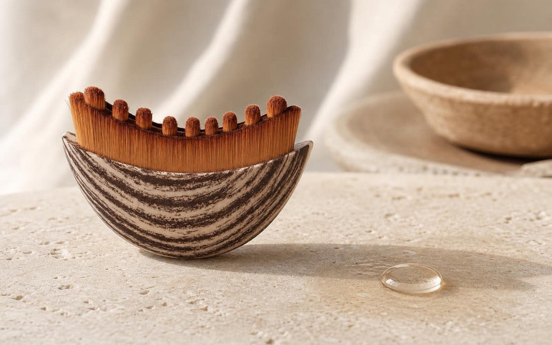 The dry bristles of the Sanamar Contour brush beside a single bead of clear facial oil on cream stone
